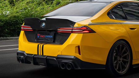 A yellow BMW 5 SERIES G60 sedan shown from the rear three-quarter angle, featuring a black carbon-fiber style rear spoiler, diffuser, and license plate frame with the TAKD CARBON logo.
