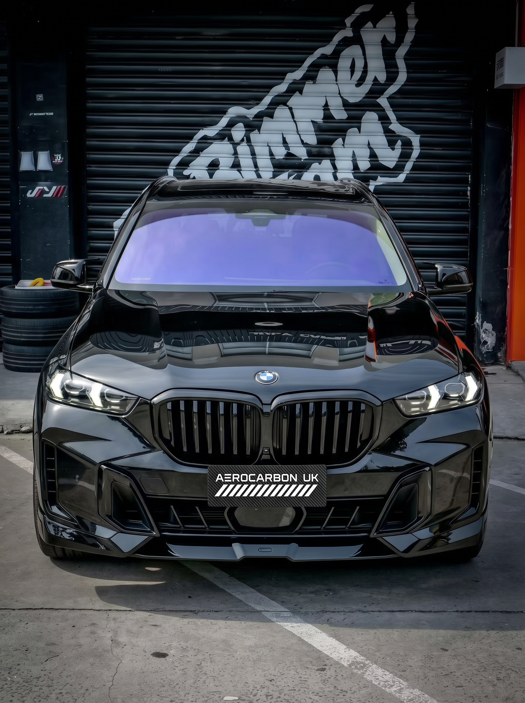 A glossy black BMW X5 G05 LCI SUV parked outdoors in front of a dark garage door with graffiti, featuring a custom front lip and a license plate that reads 'AEROCARBON UK'.