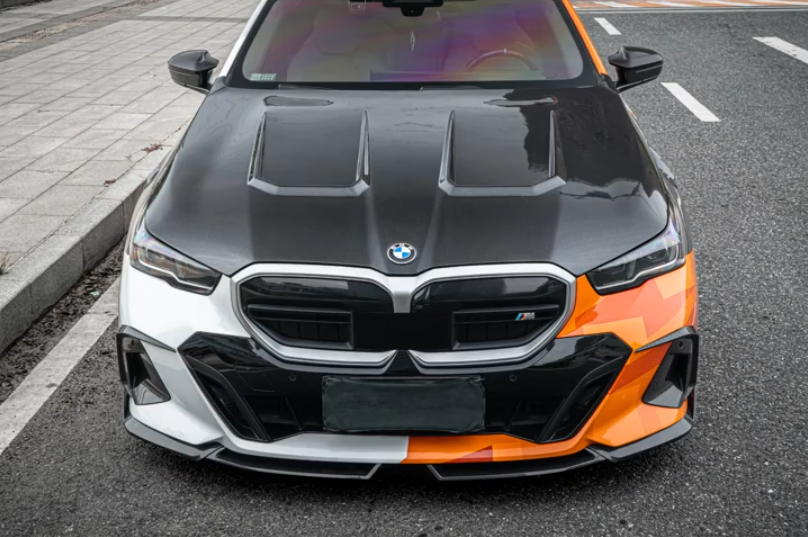 A BMW 5 SERIES sedan photographed from the front in an outdoor street setting. The car has a custom livery with a black hood, a white front bumper and lower bumper area, and a bright orange section on the right front fender and lower bumper, with the right headlight area obscured by a gray censor block.